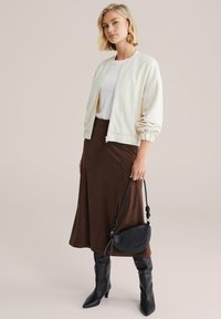 Béžová textúrovaná bomber bunda cez biely top, kombinovaná s hnedou semišovou sukňou a čiernymi čižmami nad kolená s ostrou špičkou. Malá čierna kabelka.