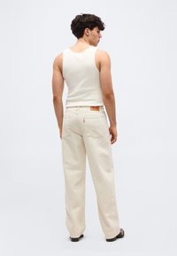 Νεαρός άνδρας με την πλάτη γυρισμένη, φοράει λευκό αμάνικο τοπ και εκρού τζιν παντελόνι με ψηλή μέση και φαρδιά πόδια, καθώς και μαύρα παπούτσια σε απλό φόντο.