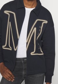 Giacca felpa blu navy con vestibilità rilassata, caratterizzata da un grande logo "M" ricamato dorato e testo, abbinata a una t-shirt bianca sottostante.