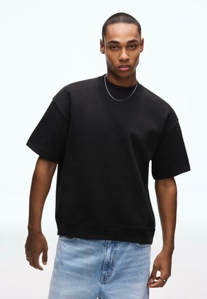 Giovane uomo con capelli corti che indossa una felpa nera oversize a maniche corte, una collana d'argento e jeans in denim azzurro chiaro su sfondo bianco.