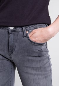 Szare dżinsy o slim fit, z przednią kieszenią i lekko przetartym materiałem na prawej udzie. Widoczny srebrny guzik i metalowe elementy.