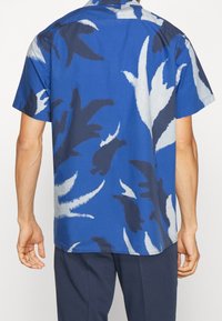 Camicia a maniche corte in tessuto blu con motivi astratti di foglie bianche e blu scuro. Presenta un colletto a camicia e un design dalla vestibilità rilassata.