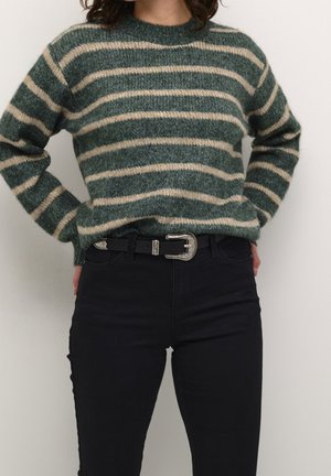 Personne portant un pull rayé vert et beige, rentré dans un pantalon noir taille haute avec une ceinture noire et une boucle argentée.