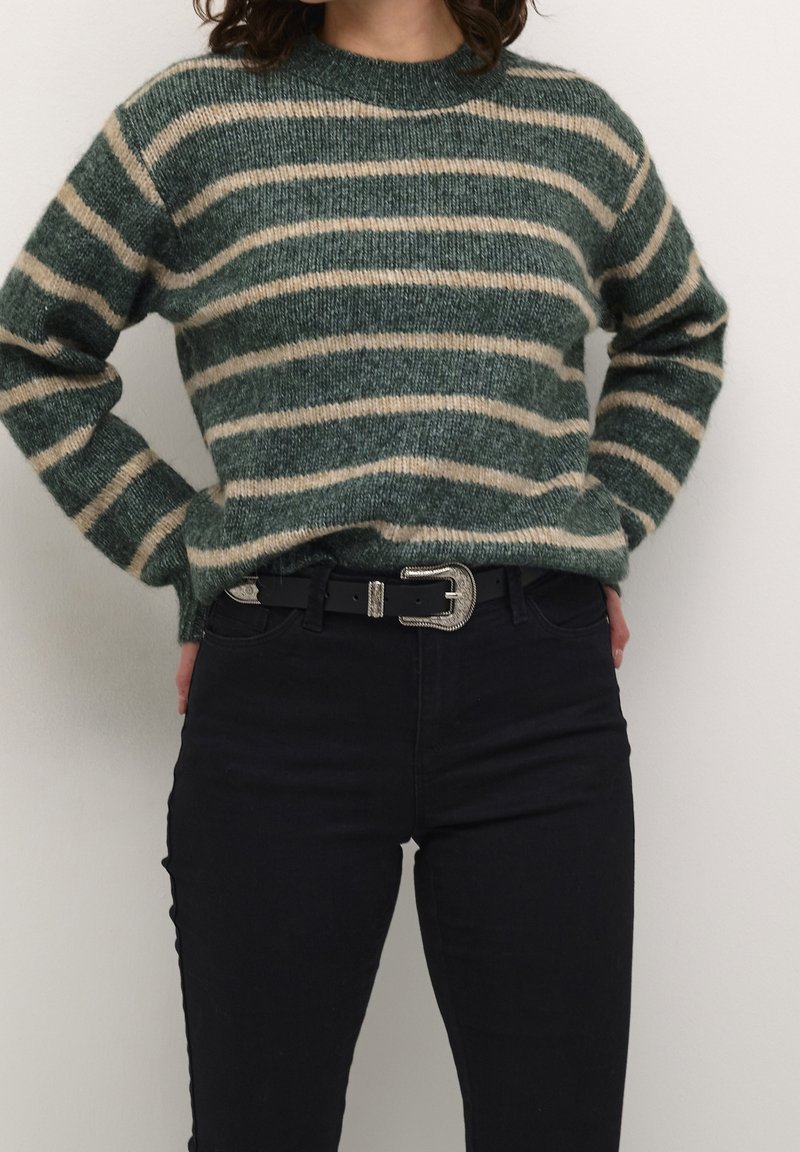 Personne portant un pull rayé vert et beige, rentré dans un pantalon noir taille haute avec une ceinture noire et une boucle argentée.