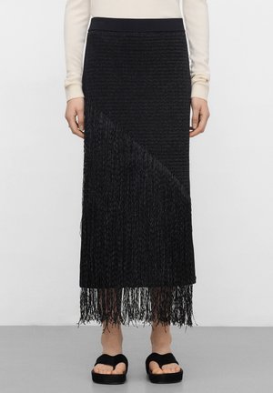 Czarna spódnica midi z teksturowanym splotem i długim frędzlowanym dołem, noszona z kremową bluzką z długim rękawem oraz czarnymi sandałami na platformie z odkrytymi palcami.