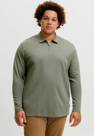 Homme aux cheveux bouclés portant un polo vert à manches longues et un pantalon marron, debout devant un fond clair uni.