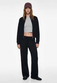 Black zip-up jacket over a gray cropped shirt, paired with black wide-leg pants. A burgundy cap accents the outfit. Smooth fabric texture throughout.