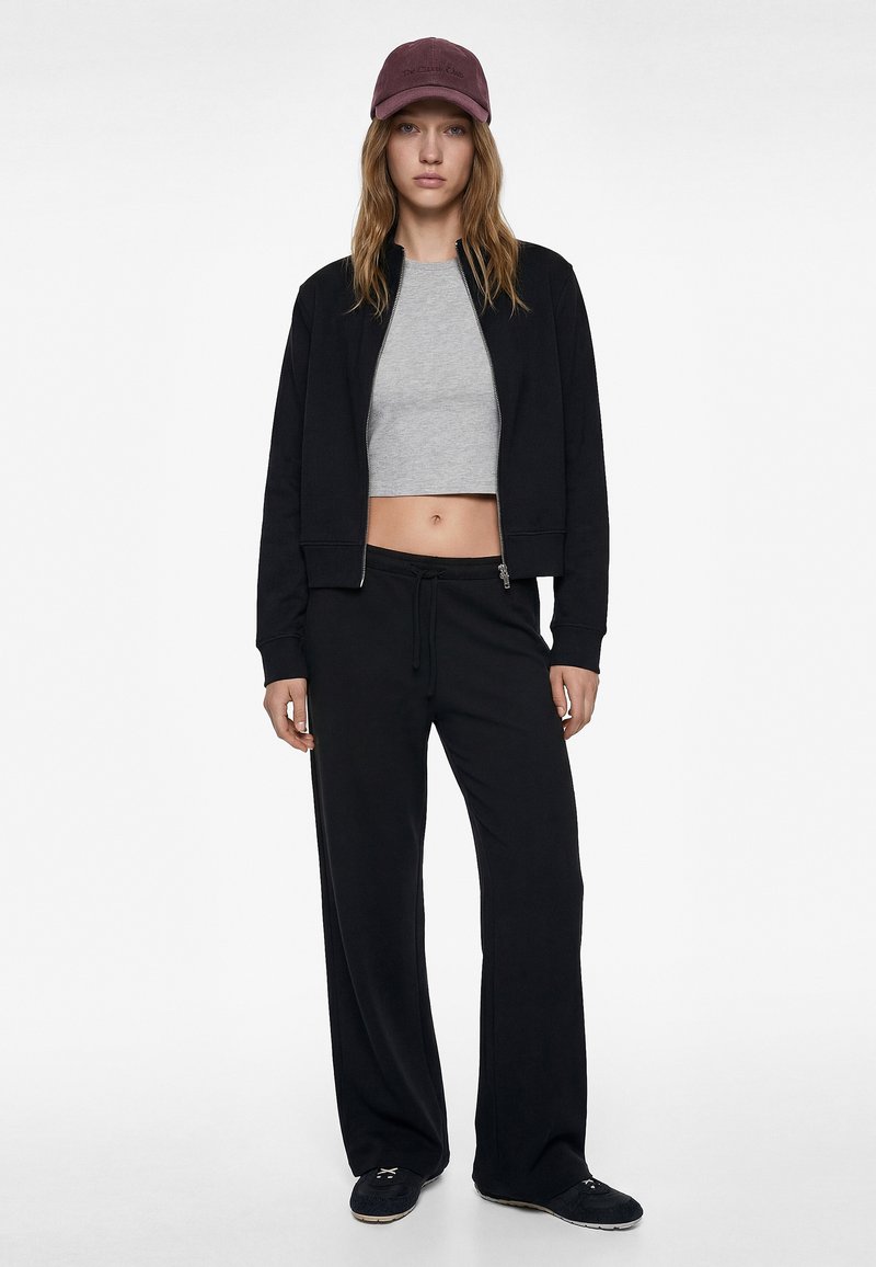 Black zip-up jacket over a gray cropped shirt, paired with black wide-leg pants. A burgundy cap accents the outfit. Smooth fabric texture throughout.