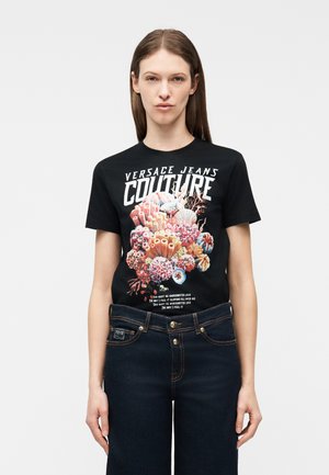 Ženska, oblečena v črno majico Versace Jeans Couture z barvnim potiskom koralnega grebena in temno modre kavbojke, stoji pred enobarvnim ozadjem.