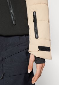 Beige Daunenjacke mit Ärmel, schwarzem Reißverschluss und Klettverschlussdetails, ausgestattet mit einem eng anliegenden Bündchen und kontrastierendem schwarzem Innenstoff. Dunkle Hose mit Logo.