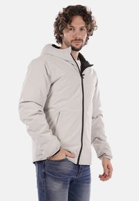 Chaqueta impermeable de color gris claro con capucha forrada en negro, cierre frontal con cremallera, puños elásticos y dos bolsillos laterales. Textura de tela suave.