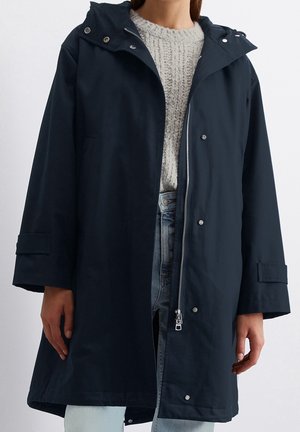 Personne portant un imperméable à capuche bleu foncé par-dessus un pull en maille gris clair et un jean bleu clair, debout devant un fond uni.