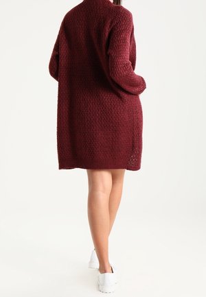 Personne portant une robe pull bordeaux texturée mi-longue et des baskets blanches, debout les mains dans les poches, tournée de dos.