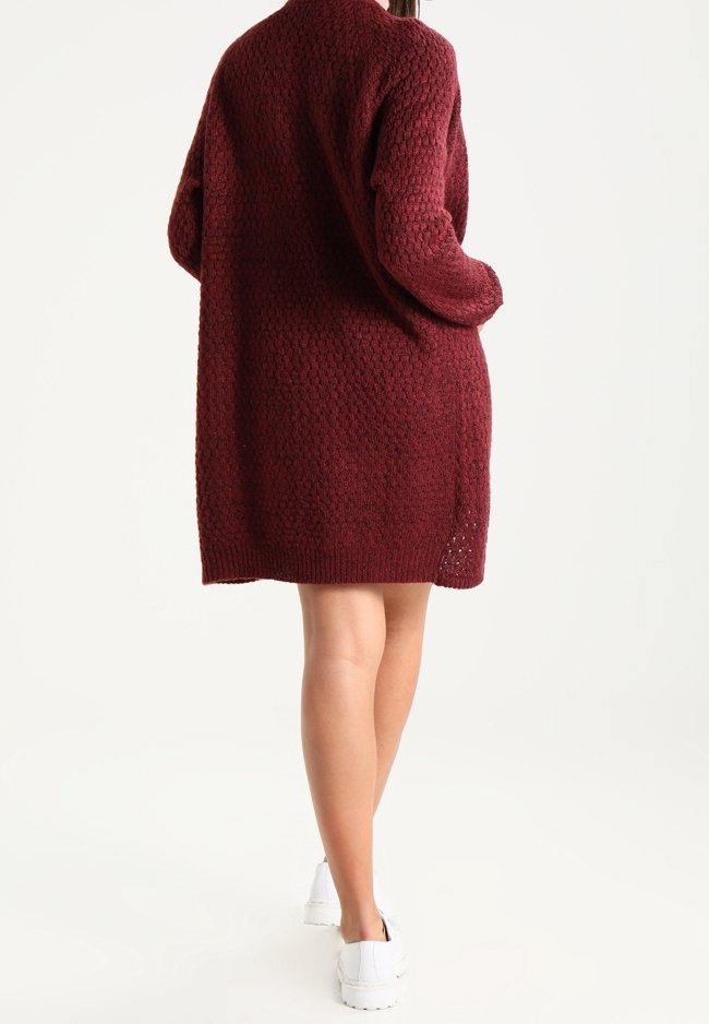 Personne portant une robe pull bordeaux texturée mi-longue et des baskets blanches, debout les mains dans les poches, tournée de dos.