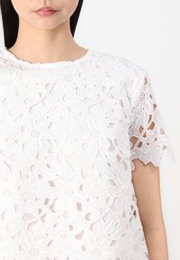 Top in pizzo floreale bianco con scollatura tonda e maniche corte. Il motivo intricato include disegni di fiori in rilievo e dettagli ricamati.