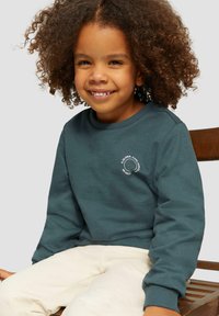 Tumši zaļš svīteris ar apaļu balto logo, apaļa kakla izgriezuma un ribotām piedurknēm. Valkāts ar gaišiem biksēm, sēžot uz koka krēsla.