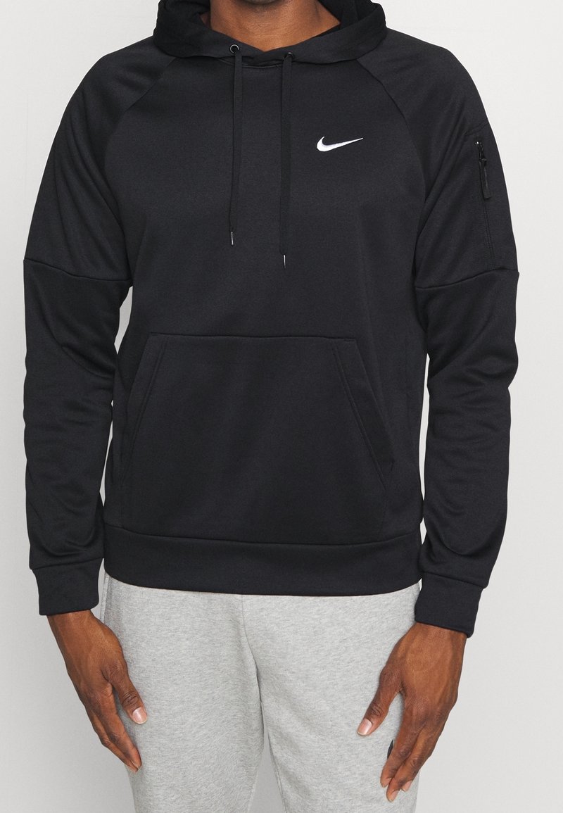 Homme portant un sweat à capuche noir Nike avec poche avant et poche zippée sur la manche, associé à un pantalon de survêtement gris clair.
