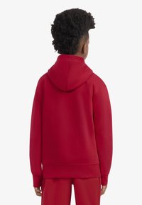 Rdeča jopa s kapuco z nizom, rebrastimi manšetami in spodnjim robom. Gladek tekstura in sproščen kroj, kombiniran z ujemajočimi rdečimi hlačami.