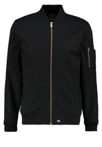 Veste bomber noire avec une fermeture éclair dorée, des poignets et une ceinture côtelés. Comprend une poche zippée sur la manche gauche et des poches latérales.