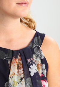 Granatowa sukienka w kwiatowy wzór, z plisowanym fasonem i miękkiego materiału. Udekorowana żywymi kwiatami w odcieniach różowego, białego i szarego.