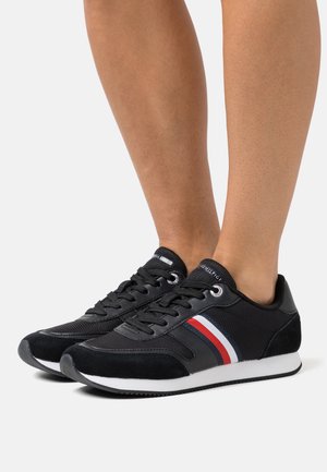 Zapatos atléticos negros con materiales de malla y ante, que presentan una suela blanca y un acento a rayas en rojo, blanco y azul.