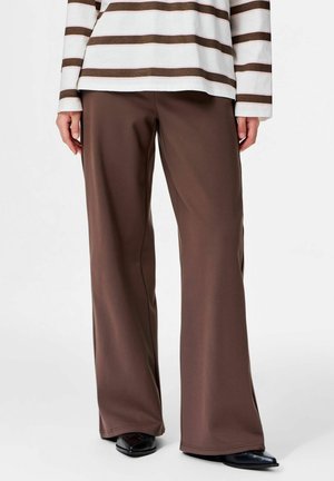 Personne portant un pantalon large marron, des chaussures noires à bout pointu, et un haut blanc à manches longues avec des rayures horizontales marron.