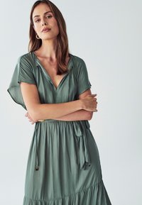 Zelené, splývavé šaty vyrobené z lehkého materiálu; mají véčkový výstřih, krátké rukávy a nařasený pas se stahovací šňůrkou. Hladká textura, délka po kolena.