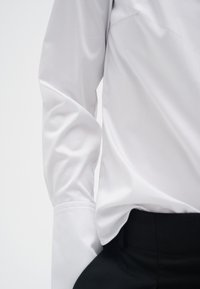 Valkoinen pitkähihainen paita puettuna korkeavyötäröisten mustien housujen sisään, vasen käsivarsi ja vyötärön alue näkyvissä yksiväristä taustaa vasten.