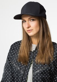Schwarze Baseballmütze kombiniert mit einer Bomberjacke im Punktmuster. Die Jacke hat einen Reißverschluss und einen strukturierten Stoff und wird über einem grauen Shirt getragen.