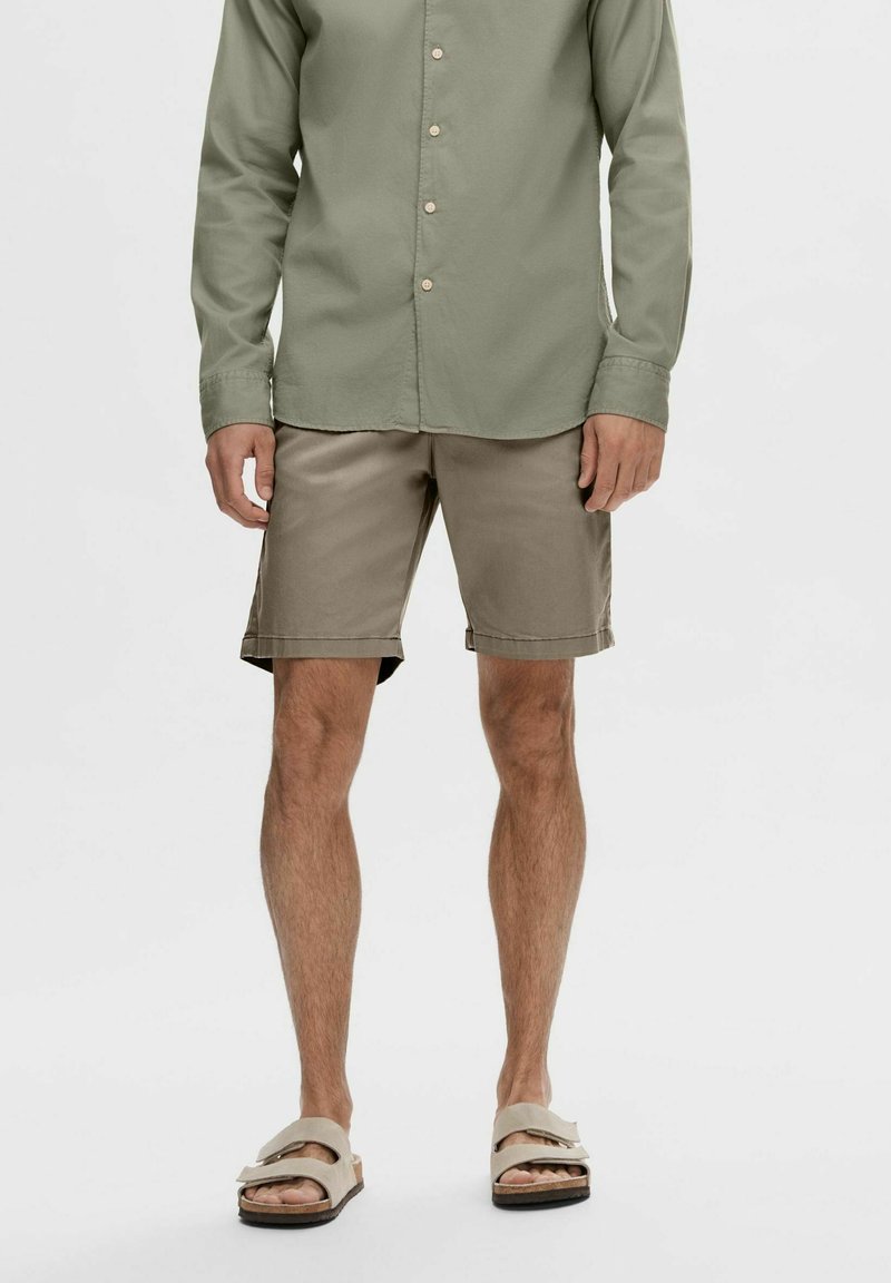 Cămașă verde măslinie cu mâneci lungi, pantaloni scurți bej și sandale bej. Pantalonii scurți au tivurile răsucite. Ținuta este casual de vară.