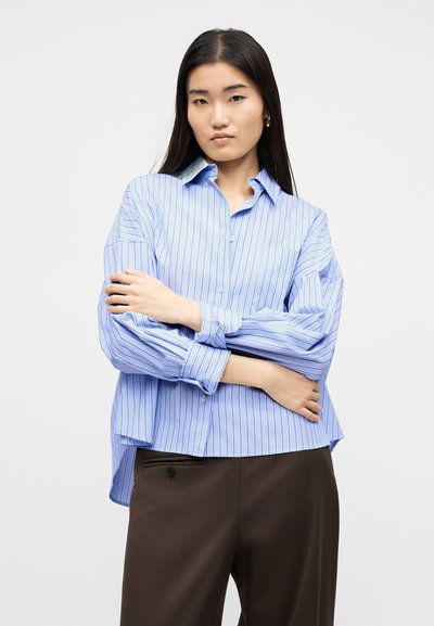 Νεαρή γυναίκα με μακριά σκούρα μαλλιά που φορά μπλε πουκάμισο με ρίγες και σκούρο καφέ παντελόνι, στέκεται με τα χέρια σταυρωμένα μπροστά σε λευκό φόντο.