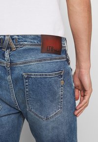 Modré denimové džíny s vybledlým omekem, se hnědou koženou etiketou označenou "LTB" a žlutým šitím. Zadní kapsy jsou viditelné.