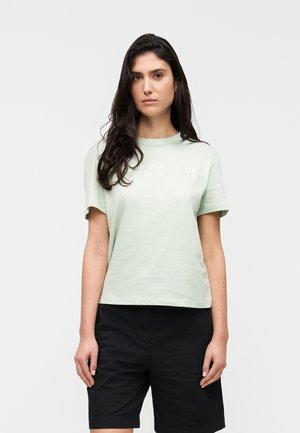 Ženska z dolgimi temnimi lasmi, ki nosi svetlo zelena kratka majica s kratkimi rokavi in črne kratke hlače do kolen, stoji na enobarvnem ozadju.