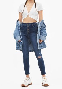Höga blå denimjeans med slitningar vid knäna. Matchat med en vit kort topp och en överdimensionerad denimjacka samt vita sneakers.