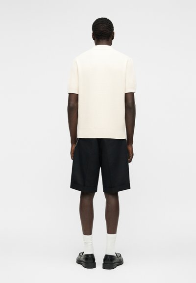 Homme portant un pull en tricot blanc à manches courtes, un short noir jusqu'aux genoux, des chaussettes blanches et des chaussures noires à boucle, debout dos au spectateur.