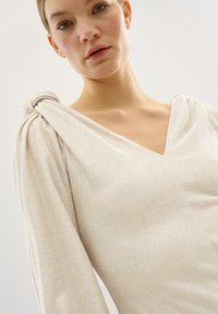 V-nyakú, hosszú ujjú felső világos bézs színben, texturált anyagból, vállán összegyűrődéses csomó részlettel. Simább felület.