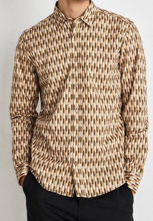 Hombre con camisa de manga larga abotonada con un patrón geométrico en beige, blanco y negro, con las manos en los bolsillos de los pantalones negros.