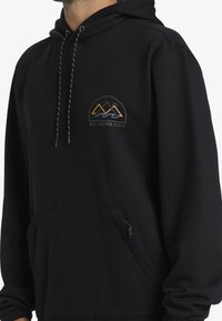 Černá mikina s kapucí vyrobená z měkkého materiálu, obsahující přední kapsu na zip a logo s horami a vlnami. Mezi detaily je také šňůrka s zlatými akcenty.