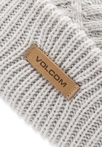 Bonnet tricoté gris avec un motif texturé et une étiquette en cuir portant le nom de la marque "VOLCOM" cousue en marron foncé.