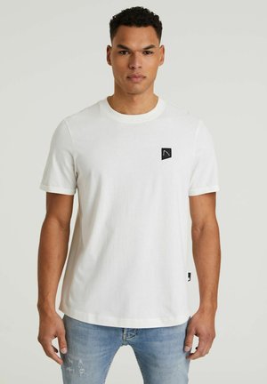 T-shirt bianco in cotone a maniche corte, con collo rotondo e una toppa nera con logo sul lato sinistro del petto. Indossato con jeans chiari strappati.