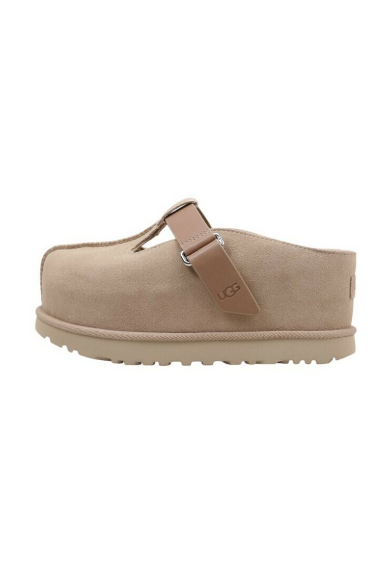Zoccoli in suede beige con una fascia e dettagli in argento, dotati di una spessa suola in gomma e punta arrotondata, progettati per comfort e facilità d'uso.