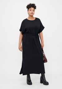 Black maxi dress with short sleeves, belted waist, side slits, and a smooth texture. Complemented by black platform boots and a burgundy bag.