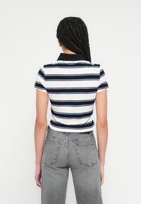 Fehér, tengerészkék és kék csíkos galléros póló rövid ujjakkal; fekete gallérral. Szürke, magas derekú farmerrel párosítva, hátulról látható.