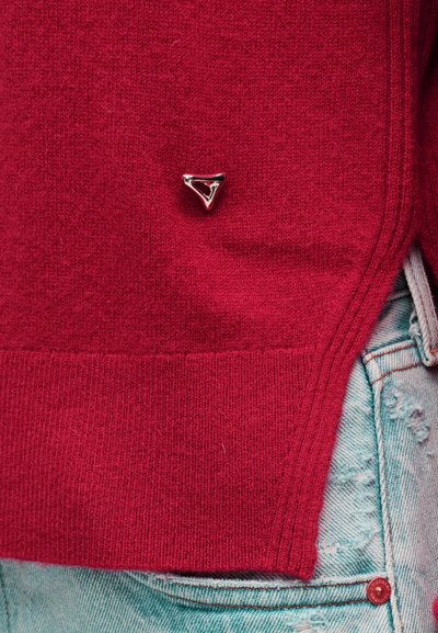 Κοντινό πλάνο του τελειώματος ενός κόκκινου πλεκτού πουλόβερ με ασημένια τριγωνική λεπτομέρεια πάνω από την τσέπη ενός ανοιχτού μπλε τζιν με φθορές.