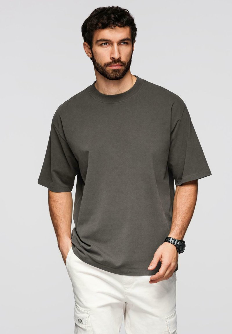 Homme barbu portant un t-shirt oversize gris foncé et un short cargo blanc, les mains dans les poches, devant un fond gris uni.