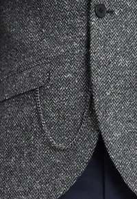 Blazer grigio a motivo herringbone realizzato con un tessuto testurizzato, dotato di un bottone nero e un dettaglio in catena argentata sulla tasca.