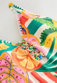 Tela de colores con estampado de frutas tropicales y hojas sobre un fondo color crema, con un detalle de tira trenzada texturizada.