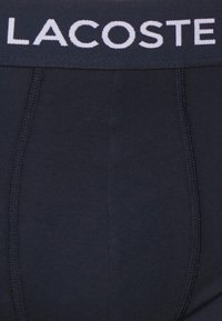 Črne bombažne boxer hlačke z širokim elastičnim pasom, na katerem je v beli barvi napis "LACOSTE". Gladka tekstura s šivanimi šivi.