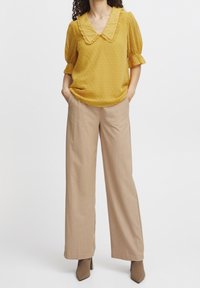 Senfgelbe Bluse mit Rüschkragen, gepunktetem Muster und Puffärmeln, kombiniert mit beigefarbenen weiten Hosen und braunen Absätzen.