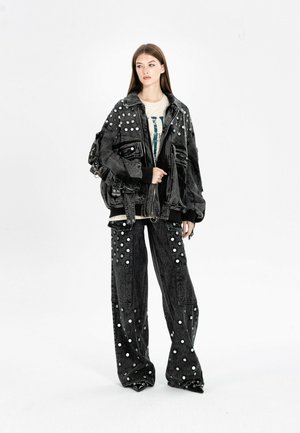 Joven mujer con chaqueta vaquera negra de gran tamaño y pantalones de pierna ancha con grandes lunares blancos, de pie sobre un fondo blanco.